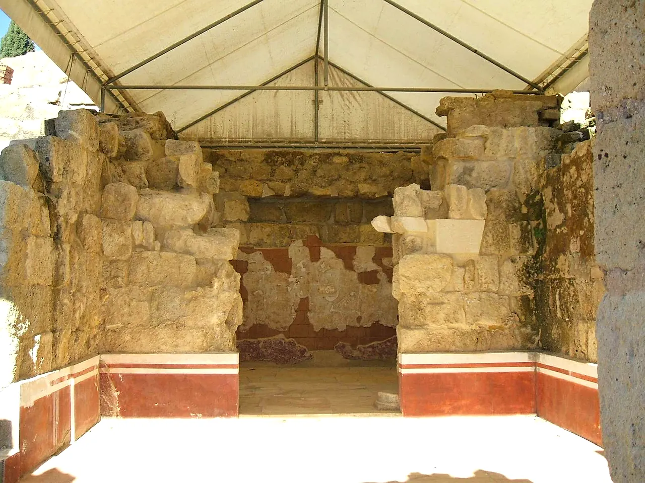 A room of Medina Azahara under a protective canopy, with stone walls and a red-painted band along the lower part of the wall